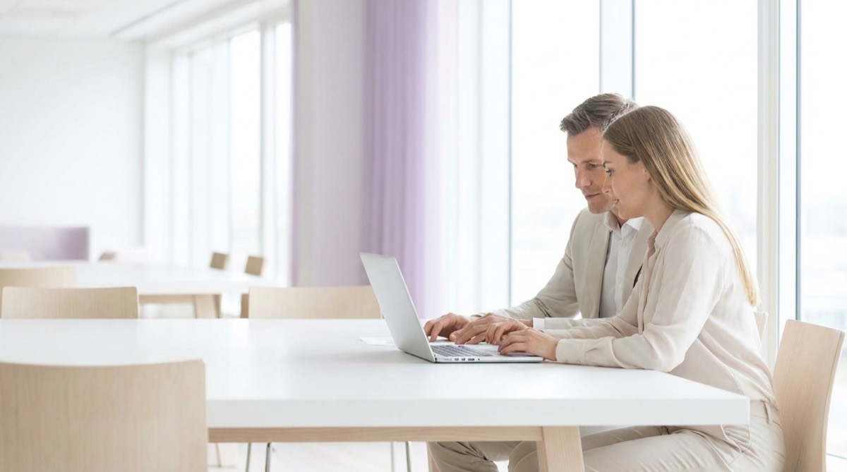 Two colleagues at a bright white table reviewing work together on a laptop.